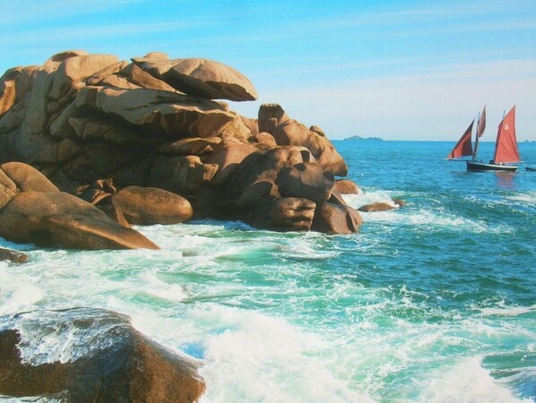 A rocky coastline with waves crashing against the rocks and a sailboat with red sails in the distance.