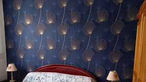 Une chambre à coucher avec une couvre-lit à motifs floraux, un papier peint bleu foncé orné de motifs de coquillages, des tables de chevet en bois avec des lampes et une armoire en bois.