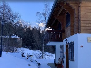 Una cabaña de madera con balcón, suelo cubierto de nieve y montañas al fondo.