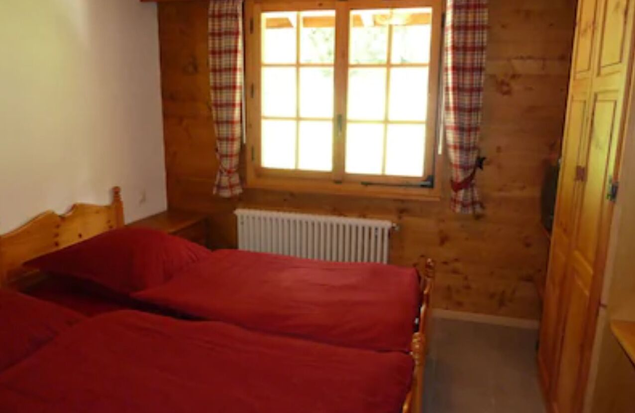 Une pièce avec un plancher en bois, un lit avec une literie rouge, une fenêtre avec des rideaux à carreaux et un radiateur.