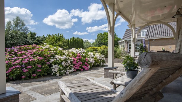 En täckt uteplats med en trästol, ett stengolv och en blomsterrabatt med rosa och vita blommor.