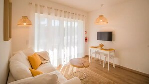 Un salon moderne avec un canapé, une petite table ronde, un téléviseur fixé au mur et une fenêtre ornée de rideaux légers.