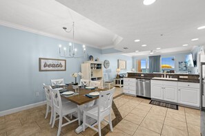 A modern kitchen with white cabinets, a dining area with a wooden table and white chairs, and a view of the beach through a window.