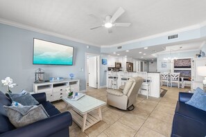 A living room with a TV, sofa, coffee table, and a chair. There is a kitchen in the background with white cabinets and a dining area.