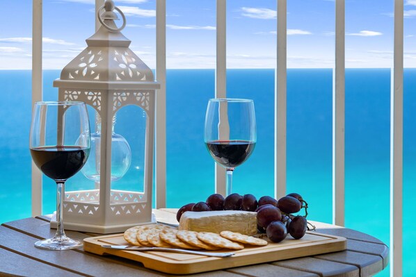 A table with a cheese, grapes, crackers, and two glasses of red wine, set against a view of the ocean.