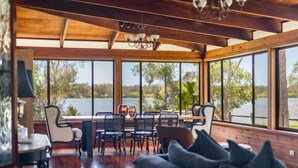 A dining area with a wooden table and chairs, large windows with a view of a lake, and a chandelier. - Riverside Ranch - Oceanstays (Yamba)
