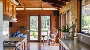 A wooden interior with a kitchen area, a seating area with a sofa and a chair, and a view of the outdoors through large glass doors. - Riverside Ranch - Oceanstays (Yamba)