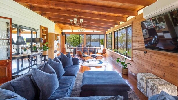 A spacious living area with a large sofa, a dining table, and a TV mounted on the wall. - Riverside Ranch - Oceanstays (Yamba)