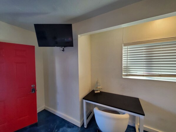 A room with a red door, a black TV mounted on the wall, a white chair, and a window with blinds.