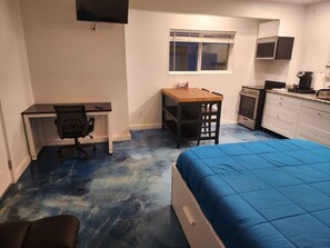 A bedroom with a bed, a desk, a chair, a small table, a window, and a kitchenette.