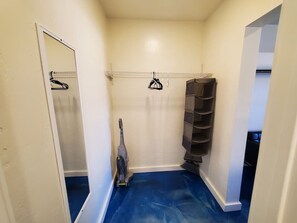 A narrow hallway with a blue floor, a vacuum cleaner, a mirror, and a rack of black shoes.