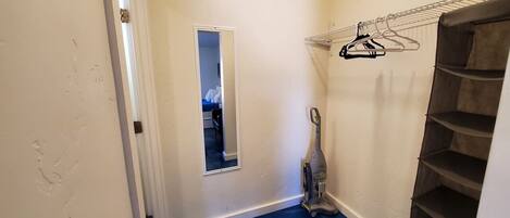 A narrow hallway with a blue floor, a mirror, a vacuum cleaner, and a shelving unit with hangers.