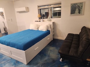A bedroom with a bed, a window, a wall-mounted air conditioner, a lamp, and a painting. - Downstairs studio on 13 st (Phoenix)