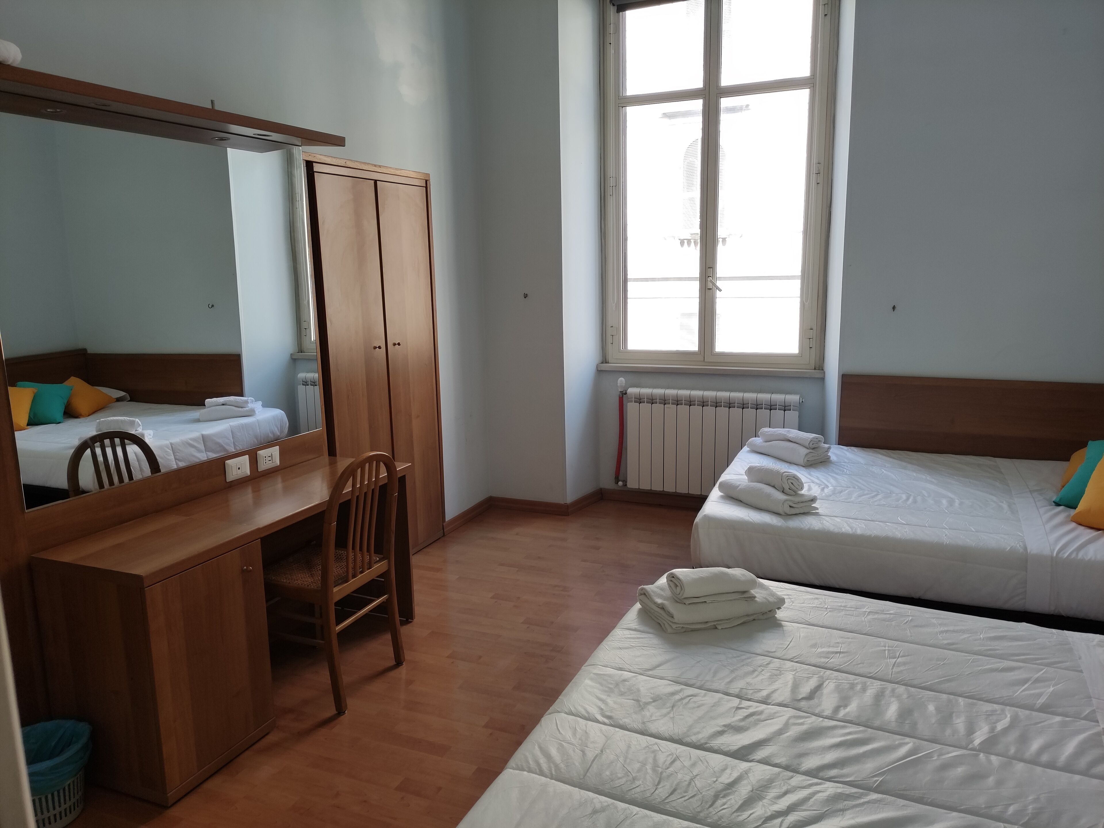 A room with two beds, a wooden desk, a chair, a radiator, and a window.