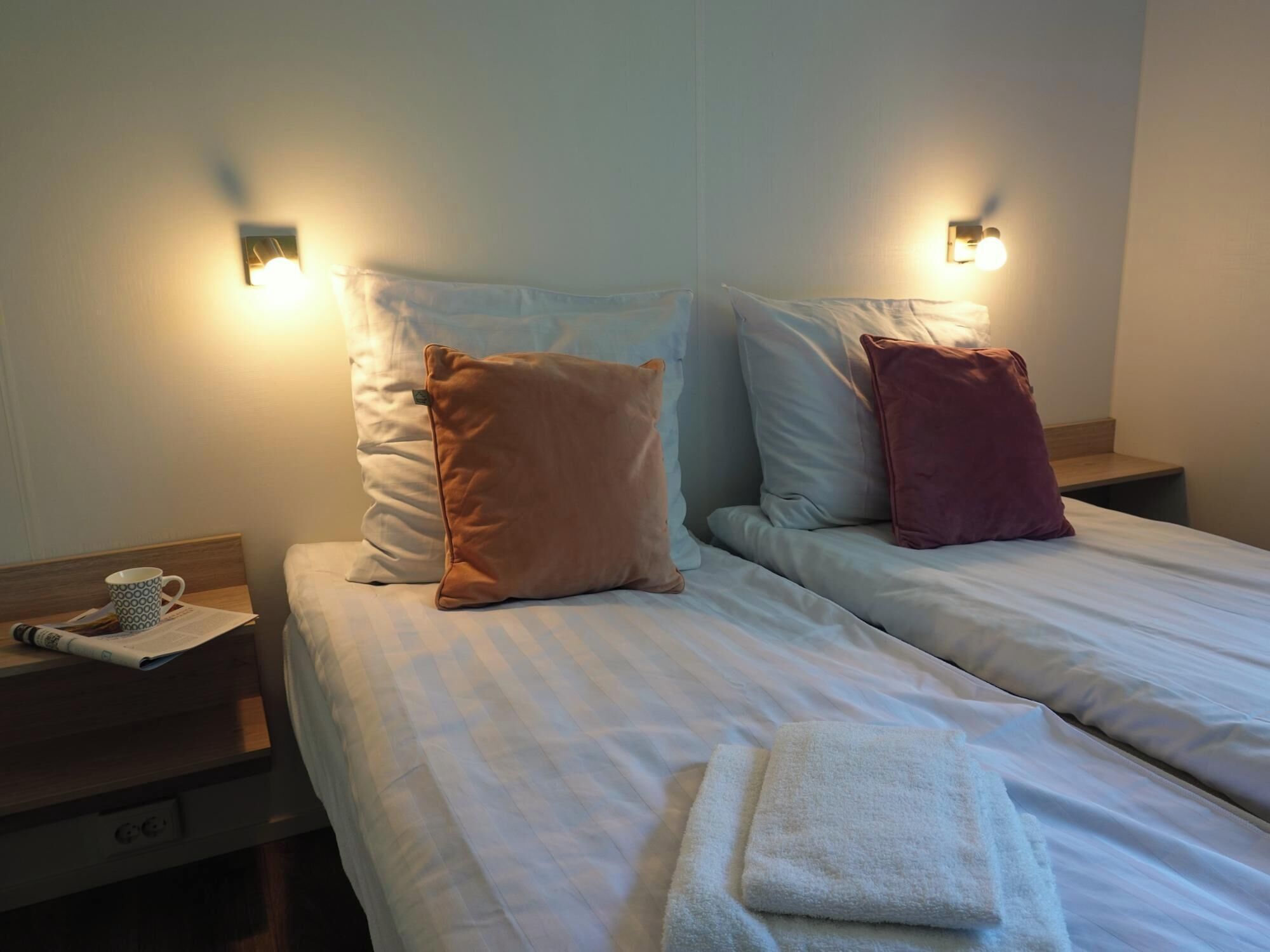 A hotel room with two beds, each with a pillow and a nightstand. There is a cup and a newspaper on the left nightstand.