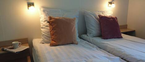 A hotel room with two beds, each with a pillow and a nightstand. There is a cup and a newspaper on the left nightstand.