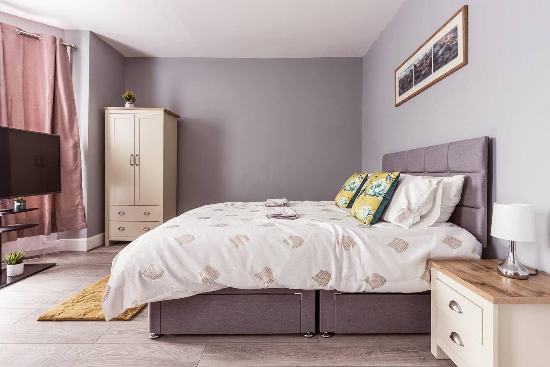 A bedroom with a bed, a nightstand, a TV, a wardrobe, and a framed picture on the wall.