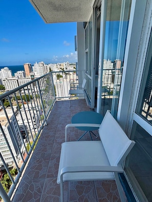 Varanda com duas cadeiras brancas e uma mesa, com vista para um horizonte urbano repleto de edifícios e céu claro.