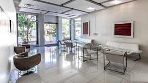 Een moderne lobby met een witte bank, een witte fauteuil en een lange bank. Er zijn grote ramen en een plant in de hoek.