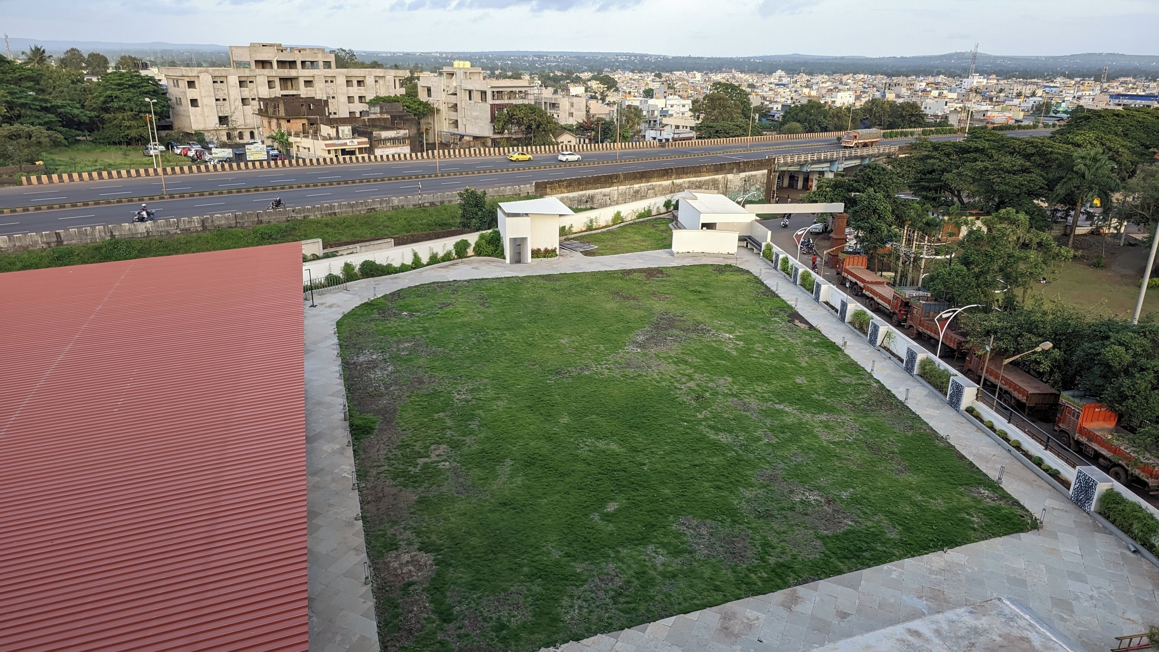 A view of a green field with a red-roofed building in the foreground, a road with vehicles, and a distant cityscape.