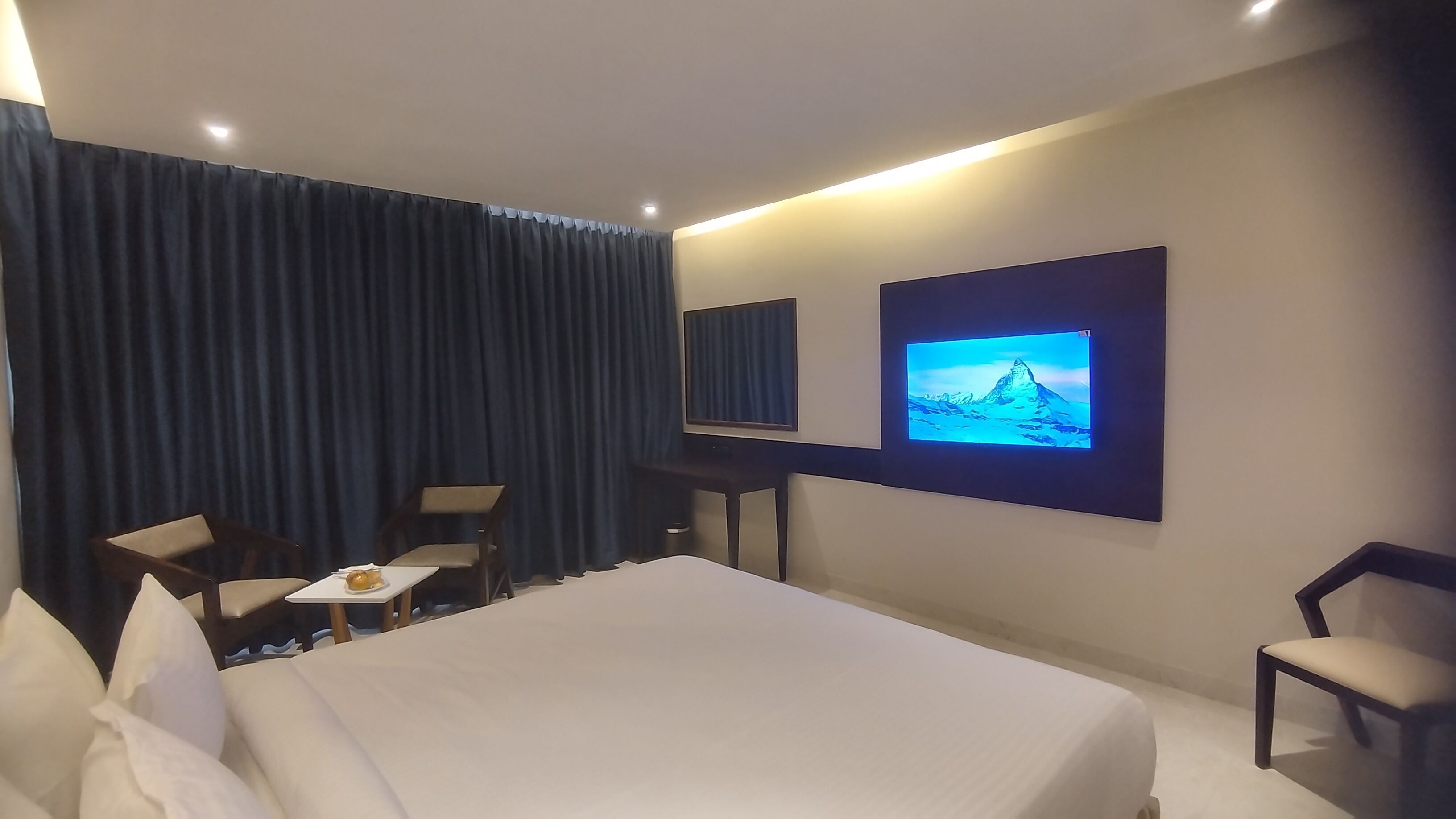 Habitación de hotel con una cama grande, un escritorio con silla, un televisor montado en la pared y un ventanal con cortinas.