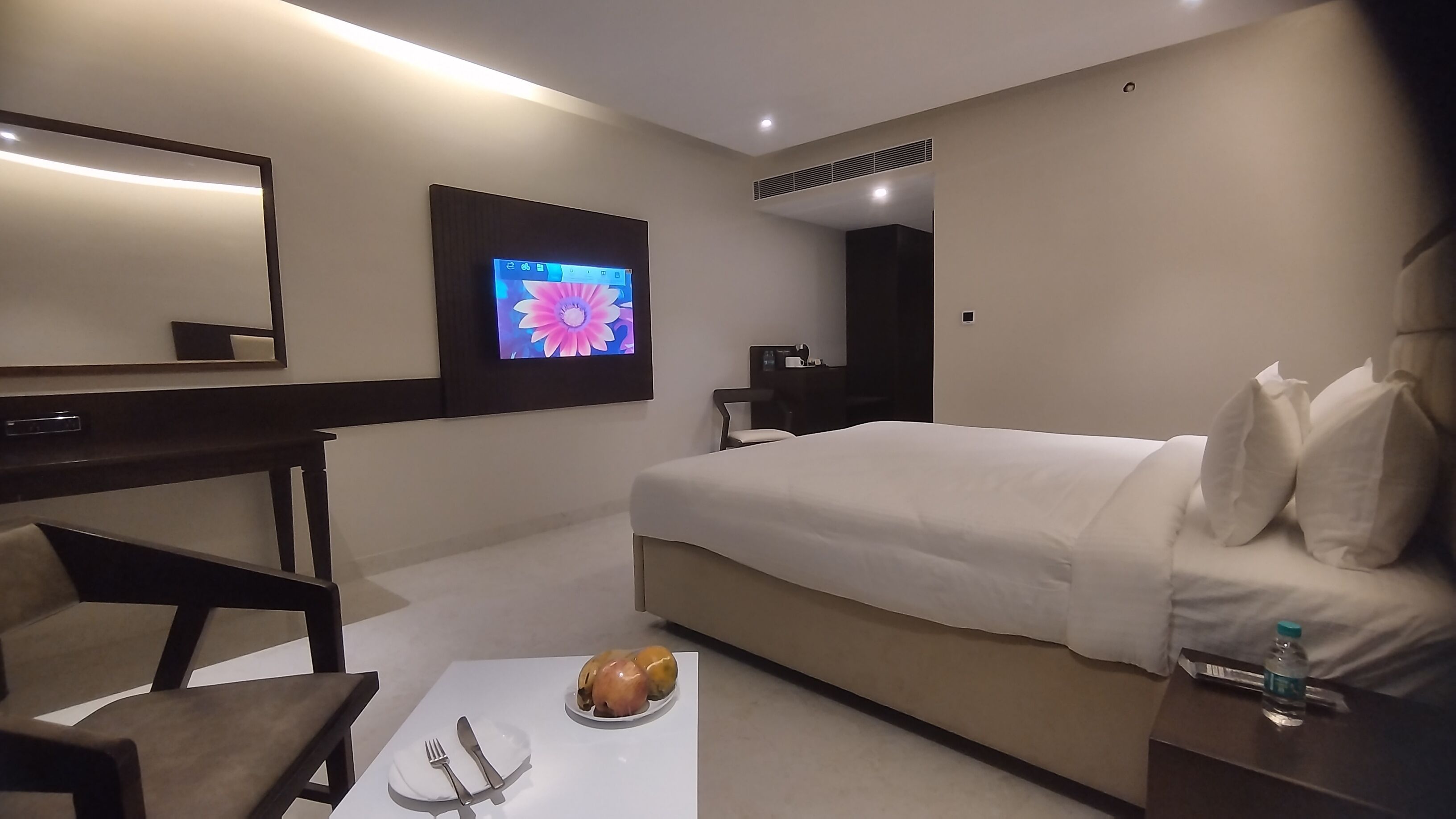 Una habitación de hotel con una cama, un escritorio, una silla, un televisor y una mesa con fruteros y una botella.