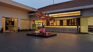 L’extérieur d’un hôtel moderne, avec une fontaine centrale, un grand arbre en pot et une entrée bien éclairée.