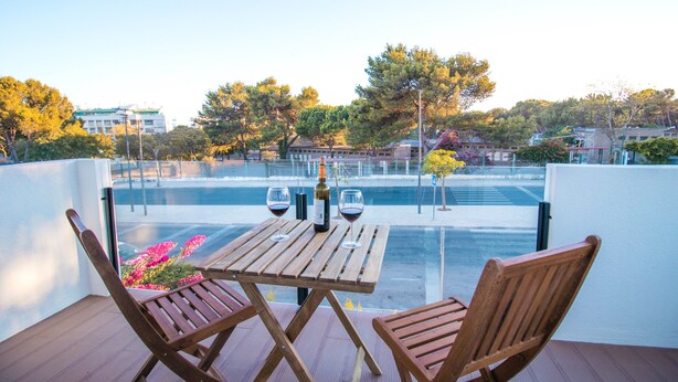 Un balcon avec une table en bois et deux chaises, donnant sur une piscine et un bâtiment, avec des arbres en arrière-plan.
