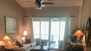 Una sala de estar con un sofá, dos sillones, una mesa de centro y un ventilador de techo. Hay un ventanal con cortinas, una planta en maceta y una lámpara sobre una mesita.