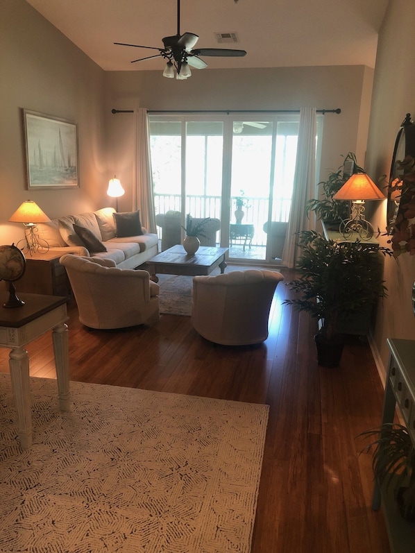 Een woonkamer met een bank, twee fauteuils, een salontafel en een plafondventilator. Er is een groot raam met gordijnen, een kamerplant en een lamp op een bijzettafeltje.