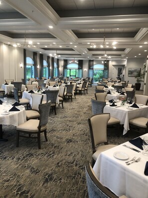 Een ruime balzaal met meerdere ronde tafels gedekt voor een diner, met witte tafelkleden en donkere stoelen.