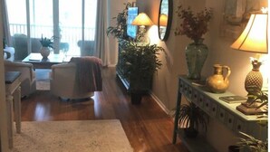Una sala de estar con piso de madera, alfombra blanca, sofá, mesa de centro y un comedor con mesa y sillas. Hay plantas en macetas y una lámpara sobre una consola.