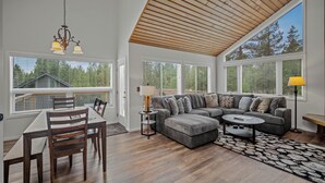 Ein geräumiges Wohnzimmer mit Holzbalkendecke, großen Fenstern, einem grauen Ecksofa, einem Esstisch mit Stühlen und einem Couchtisch.