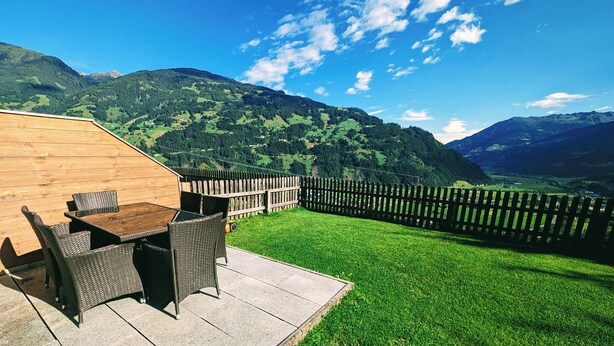 Pátio com móveis de vime e mesa de madeira, com vista para um gramado verdejante e montanhas.