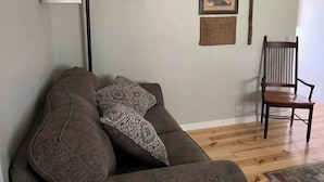 A living room with a brown sofa, a patterned throw pillow, a tall floor lamp, a framed picture, a woven placemat, and a wooden chair.