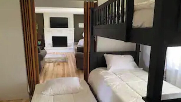 A room with two bunk beds, a wooden floor, a fireplace, and a view of a living area.