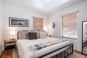 Une chambre à coucher comprenant un lit, des tables de chevet, une commode, une fenêtre avec des stores et un tableau encadré accroché au mur.