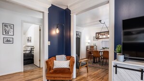 A modern living room with a dark blue accent wall, a wooden floor, a brown armchair, a dining area with a chandelier, and a flat-screen TV.
