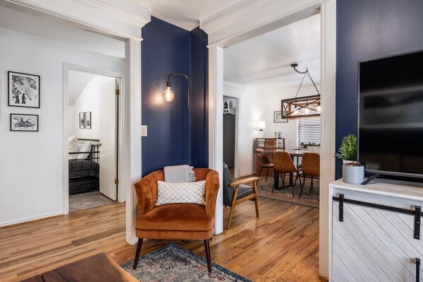 Un salon moderne avec un mur d’accent bleu foncé, un plancher en bois, un fauteuil marron, un coin repas agrémenté d’un lustre et un téléviseur à écran plat.