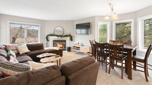 A spacious living room with a fireplace, a dining area, and large windows offering a view of the outdoors. - Mount Snow Condo: 4 min Walk to Sundance Lift (High Speed - 6 Seat) (Dover)
