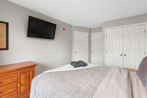 Une chambre à coucher avec un lit, un téléviseur fixé au mur, une commode en bois et une porte à deux battants peinte en blanc.