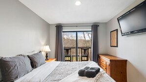 A bedroom with a large bed, a dresser, a TV mounted on the wall, and a balcony with a view of trees. - Mount Snow Condo: 4 min Walk to Sundance Lift (High Speed - 6 Seat) (Dover)