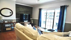 Ein Wohnzimmer mit einem beigefarbenen Sofa, einem Flachbildfernseher, einem Holztisch und einem großen Fenster mit blauen Vorhängen.