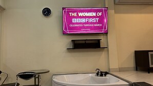 A modern bathroom with a freestanding bathtub, a wall-mounted TV displaying 'The Women of BBC First', a clock, and a small table with chairs. - Freeway Pattay make you feel as home (Pattaya)