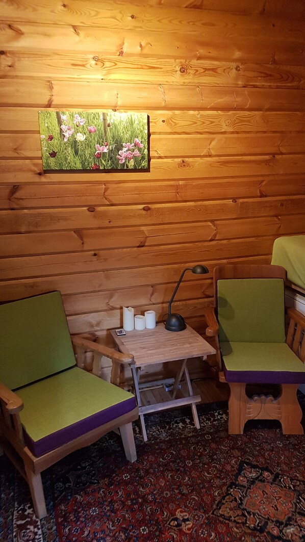 A cozy cabin room with wooden walls, two green cushioned chairs, a small wooden table with a lamp, and a painting of flowers. - "Forest Bathing" in Wendland, near the Elbe—hiking and biking right on your doorstep (Hitzacker (Elbe))