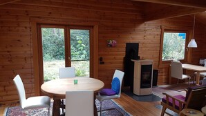 Innenansicht einer Holzhütte mit einem runden Esstisch, weißen Stühlen, einem Kamin und großen Fenstern mit Blick auf Grünfläche.