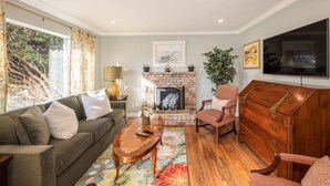 Una sala de estar con sofá, sillón, mesa de centro de madera, chimenea y televisor montado en la pared.