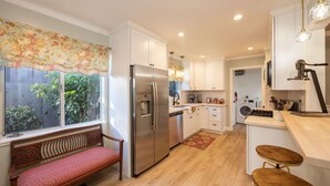 Cocina moderna con gabinetes blancos, refrigerador de acero inoxidable, lavavajillas integrado y un banco de madera junto a la ventana.