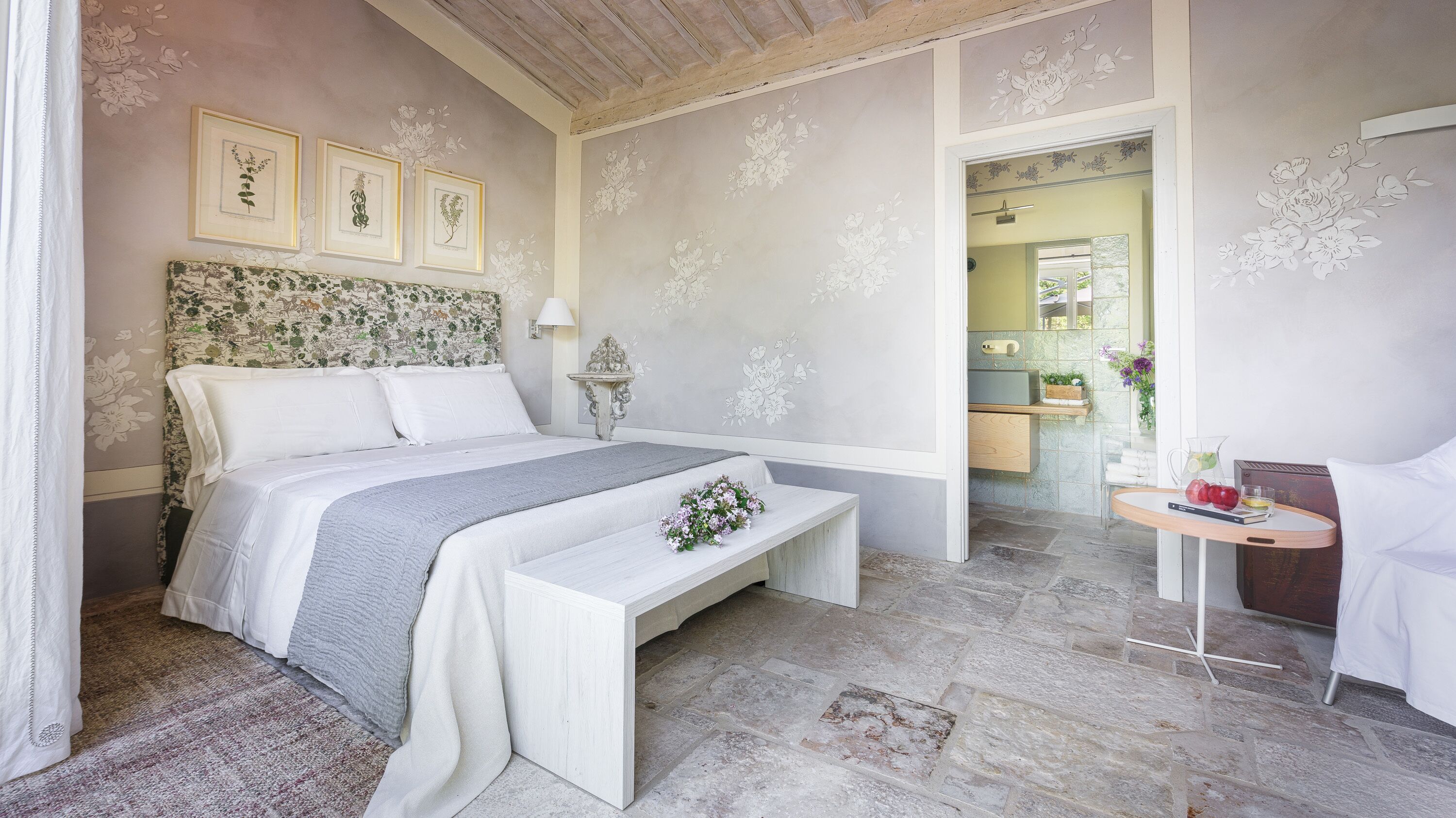 A bedroom with a large bed, a bedside table, a bench, and a small table with a lamp. There are framed botanical prints on the wall and a door leading to a bathroom.