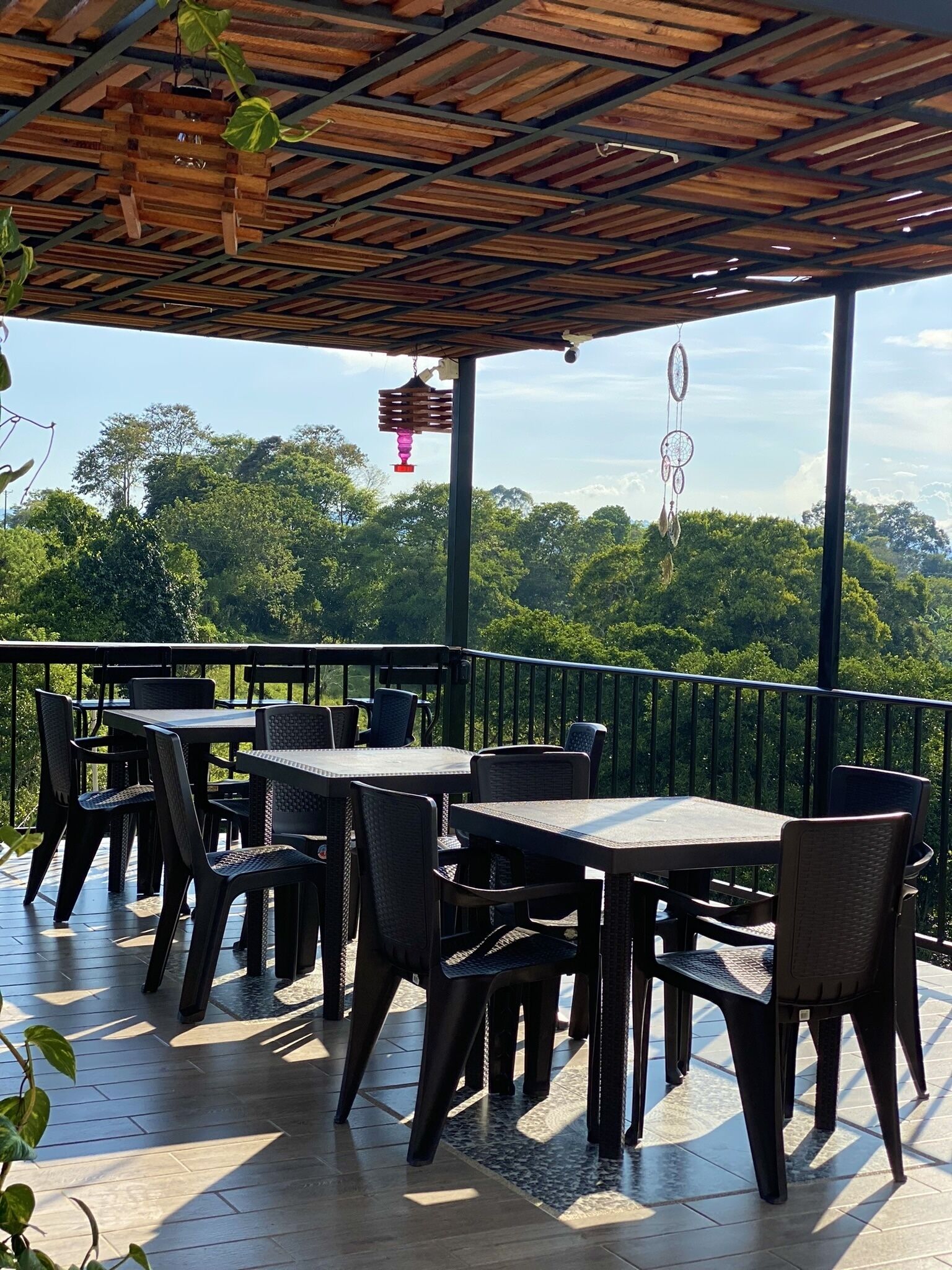 An outdoor patio with tables and chairs, a wooden ceiling, and a view of a forested landscape.